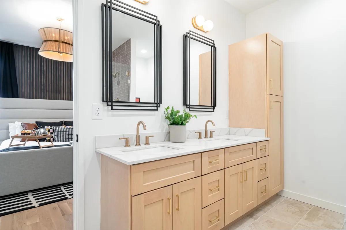 A stylish double vanity bathroom featuring gold faucets, framed mirrors, potted plants, and abundant storage cabinets, set in a calm and serene atmosphere. The space is organized and designed for both function and relaxation.