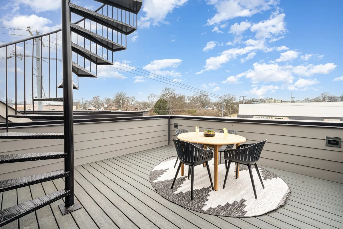 This image captures a rooftop terrace at sunset, featuring a spiral staircase, a dining table with chairs, and a decorative rug. The city skyline glows in the background, and the setup invites both social gatherings and peaceful meals under the open sky.