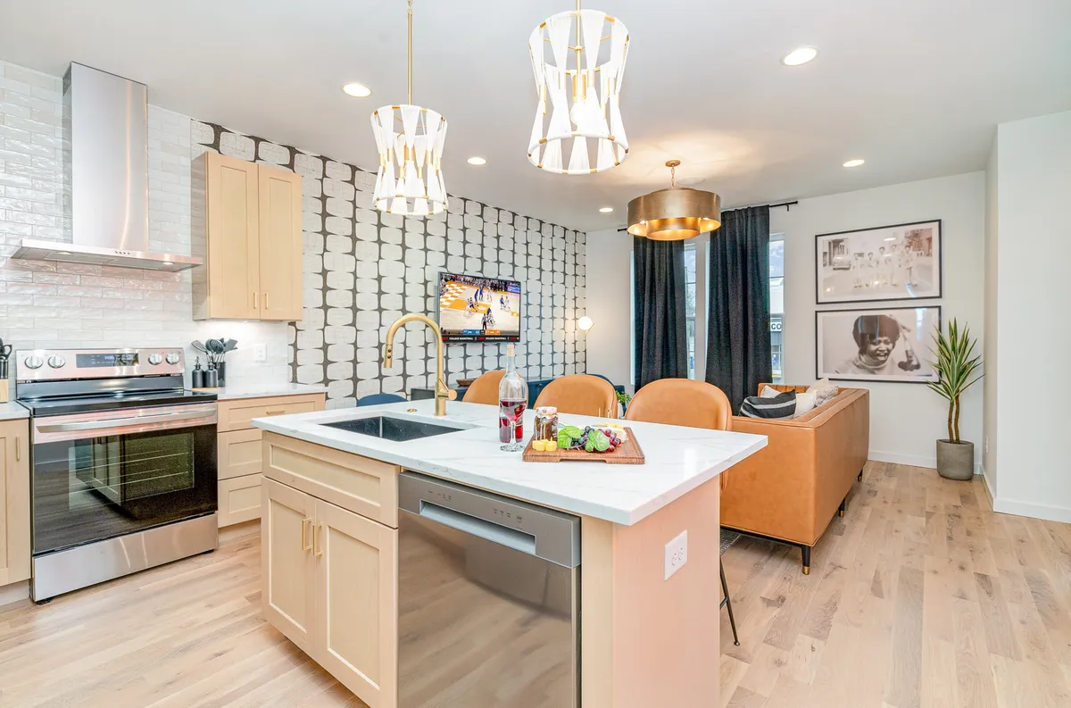 A bright, welcoming open-plan space featuring a central kitchen island with bar seating, stylish decorative light fixtures hanging overhead, modern wall art, and a seamless transition to a comfortable seating area. The setup is ideal for both relaxed gatherings and social events.