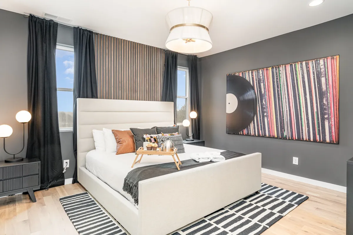 A cozy bedroom scene with a bed adorned with decorative pillows, large wall art depicting a vinyl record, and stylish bedside lamps. The room is inviting and designed for relaxation and comfort.