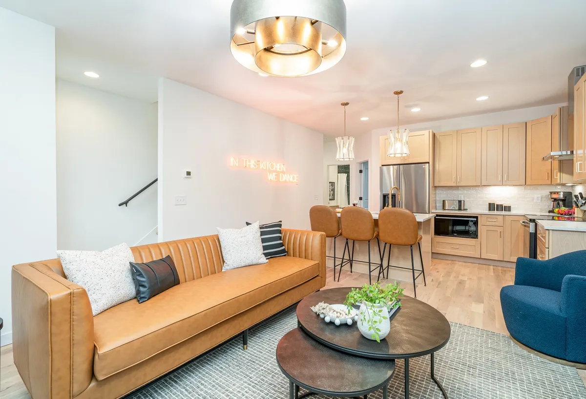 The image showcases a stylish, open-plan living area featuring a plush leather sofa, a modern kitchen island with seating, chic decorative lighting, and vibrant artwork on the wall. The scene evokes an inviting atmosphere perfect for social gatherings, cooking, and relaxing.
