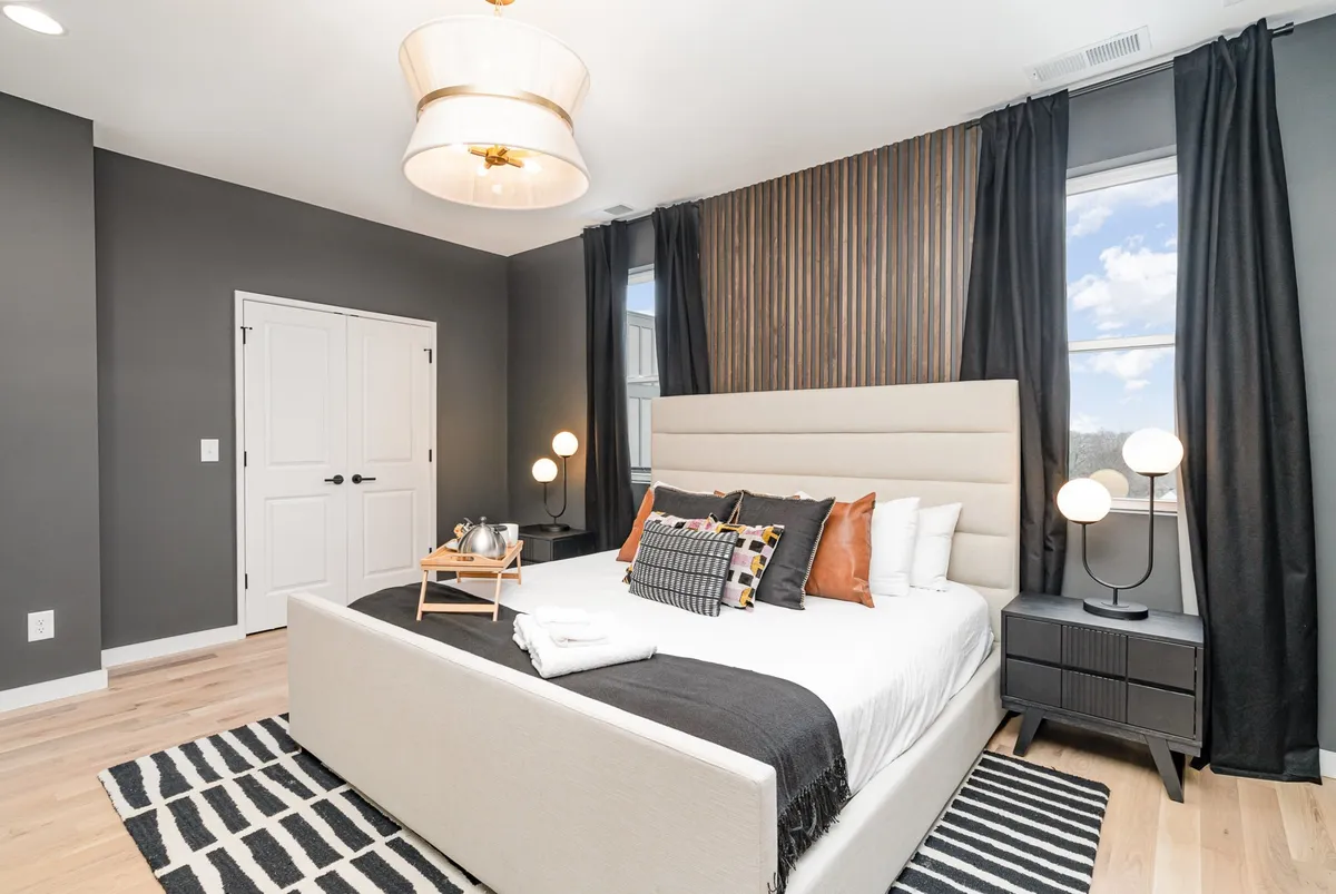 A serene and welcoming bedroom featuring a luxurious bed with decorative pillows, nightstands with modern lamps, and a chic light fixture. The space feels restful and inviting, perfect for relaxation after a long day.